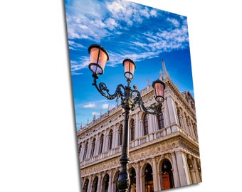Puede incluir: Una farola negra adornada con tres luces, dos de las cuales están encendidas, se encuentra frente a un gran edificio blanco con ventanas arqueadas y un cielo azul con nubes blancas en el fondo.