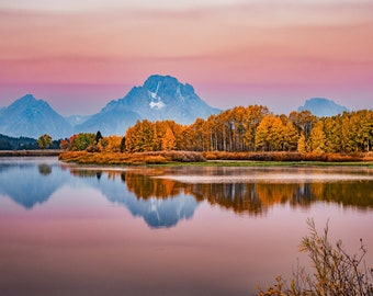 Puede incluir: Una vista panorámica de una cordillera reflejada en un lago tranquilo. Las montañas son de un suave color azul y el lago es de un rosa pálido. En primer plano, una línea de árboles con hojas amarillas crea una escena otoñal vibrante.