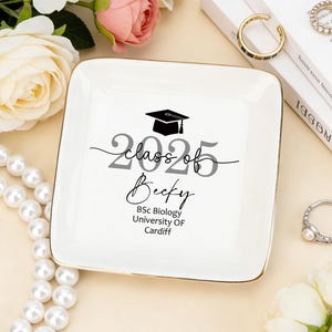 Może przedstawiać: Kwadratowa, biała ceramiczna patera na biżuterię ze złotą obwódką. Patera zawiera napis "Class of 2025 Becky" z grafiką czapki absolwenta. Poniżej znajduje się napis "BSc Biology University OF Cardiff". Patera jest otoczona biżuterią i kwiatami.
