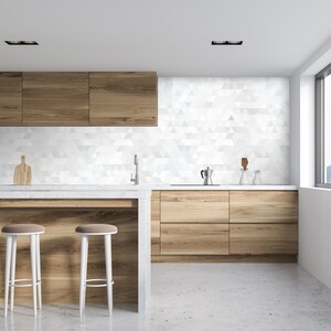 Puede incluir: Una cocina moderna con gabinetes de madera clara, una encimera blanca y un salpicadero de azulejos geométricos. Dos taburetes blancos con asientos marrones están frente a una isla de madera. Se ven un grifo de acero inoxidable y una cafetera.