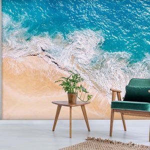 Puede incluir: Un sillón de terciopelo verde con patas de madera delante de un mural de pared que representa una vista aérea de una playa de arena y olas de color turquesa.
