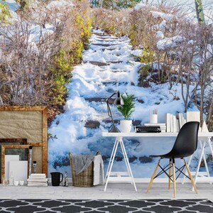 Pode incluir: Um caminho nevado com degraus de pedra que sobem uma colina. O caminho é ladeado por galhos nus e arbustos cobertos de neve. Uma mesa branca com uma cadeira preta está em primeiro plano, juntamente com uma lâmpada, uma planta e uma cesta.