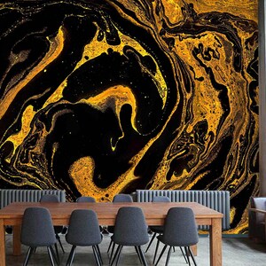 May include: Abstract wall art featuring a swirling pattern of gold and black. The design resembles marble or liquid paint, with flowing lines and contrasting colors. The artwork is displayed on a wall in a room with a wooden table and chairs.