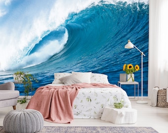 Puede incluir: Un dormitorio con un gran mural de pared de una ola azul del océano rompiendo. La cama tiene un edredón floral blanco y verde y una manta rosa. Hay un reposapiés de punto gris y una mesita de noche blanca con un cuenco de fruta verde.