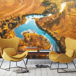 Puede incluir: Dos sillones amarillos con marcos de metal negro se encuentran frente a una mesa de café con libros apilados sobre ella. Las sillas dan a una pared con un mural fotográfico de un río sinuoso a través de un campo de hierba dorada.