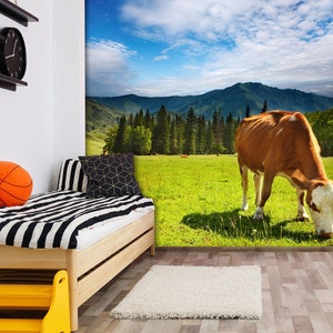 May include: A brown and white cow grazes in a green field with mountains in the background. The sky is blue with white clouds.