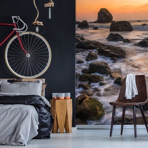 May include: A bedroom interior with a black wall, a bed with gray and black bedding, a wooden nightstand with three copper candle holders, a brown leather chair with a white towel draped over the back, and a wall mural depicting a rocky beach at sunset.