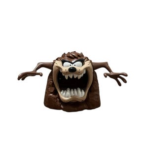May include: A brown Taz figure with an open mouth and outstretched arms. The cartoon character has a tan face, white teeth, and black eyes. The figure is a collectible item from the Looney Tunes franchise.