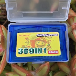 以下が含まれることがあります： 青いゲームボーイアドバンスのカートリッジケースで、開くと「369IN1」と「2048M」の文字が入ったゲームカートリッジが現れます。カートリッジには漫画のキャラクターが描かれた黄色のラベルが付いています。ケースは緑とオレンジの多肉植物に囲まれています。