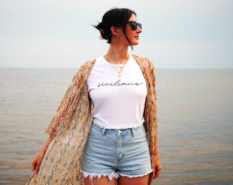 May include: A woman wearing a white t-shirt with the word "siciliana" written in black script, a brown and beige patterned kimono, and denim shorts. She is standing on a beach with the ocean in the background.
