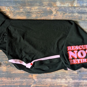 May include: Black dog shirt with the text "Rescued Not Retired" in pink and red. The shirt has a pink trim and a folded collar. The shirt is laying on a wooden surface.