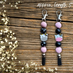 Puede incluir: Dos llaveros de vaca de color rosa y negro con borlas negras. Cada llavero tiene un anillo de llave plateado y mide aproximadamente 12,7 cm de largo.