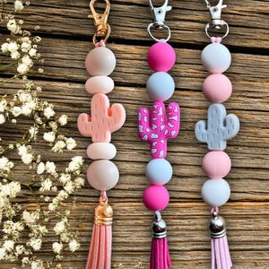 Puede incluir: Tres llaveros de cuentas con dijes de cactus. Los llaveros presentan una variedad de cuentas de colores pastel, incluyendo rosa, blanco y azul. Cada llavero tiene un dije de cactus de diferente color y una borla. Los llaveros se exhiben sobre una superficie de madera.