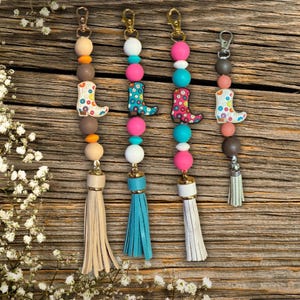 May include: Four colorful keychains with a boot charm, beads, and a tassel. The keychains are brown, pink, turquoise, and white.