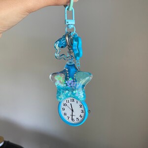 May include: A blue quartz watch with a star-shaped pendant filled with blue glitter and small stars. The watch has a white face with black numbers and hands. The keychain has a blue clasp and several charms.