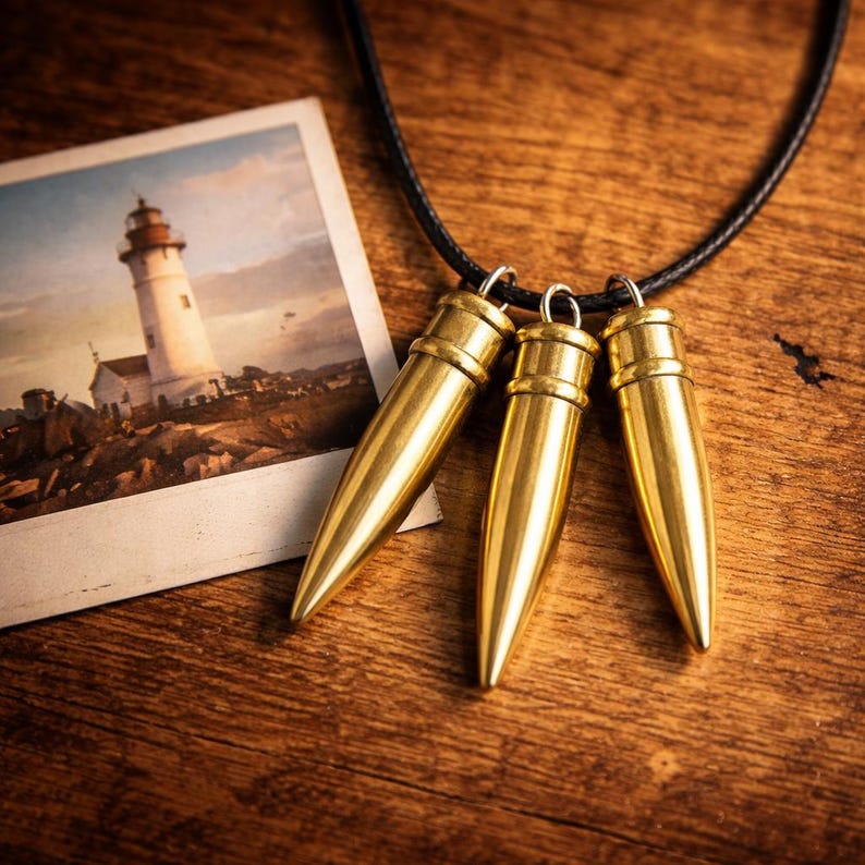 Pu&ograve; includere: Una collana con tre ciondoli dorati a forma di proiettile appesi a un cordino nero. Sullo sfondo una fotografia vintage di un faro, il tutto appoggiato su una superficie di legno. I ciondoli hanno una lucentezza metallica.