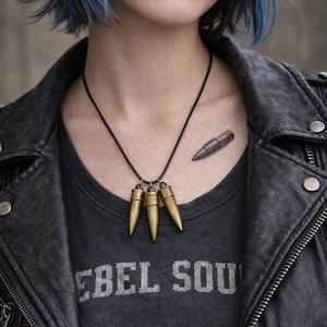 Pu&ograve; includere: Una collana con cordino nero con tre ciondoli a forma di proiettile color oro. La persona indossa una maglietta grigia con la scritta "REBEL SOUL" e una giacca di pelle nera. Un tatuaggio a forma di proiettile &egrave; visibile sul petto.