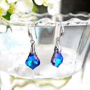 May include: A pair of silver-toned dangle earrings with teardrop-shaped, faceted blue and purple crystals. The earrings are suspended from a simple hook and are displayed in a clear glass bowl.