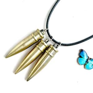 Pu&ograve; includere: Una collana con tre ciondoli a forma di proiettile color oro sospesi a un cordino nero. &Egrave; visibile anche un piccolo ciondolo a farfalla blu. I ciondoli hanno una finitura metallica e sono disposti a triangolo.