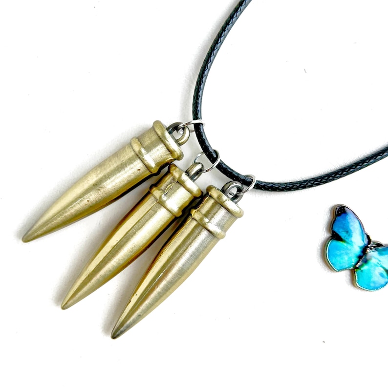 Pu&ograve; includere: Una collana con tre ciondoli a forma di proiettile color oro sospesi a un cordino nero. &Egrave; visibile anche un piccolo ciondolo a farfalla blu. I ciondoli hanno una finitura metallica e sono disposti a triangolo.