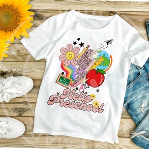Puede incluir: Camiseta blanca con un gráfico rosa de "Hello Preschool". El gráfico incluye un arcoíris, una flor, un lápiz, una regla, tijeras, una manzana, un globo terráqueo y una pizarra con las letras ABC.