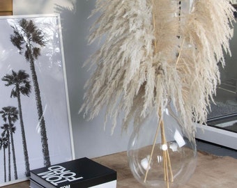 May include: A clear glass vase with a wide base and a narrow neck, filled with dried pampas grass. The vase is sitting on a wooden table next to a black book with white text that reads "TOM FORD CELINE".