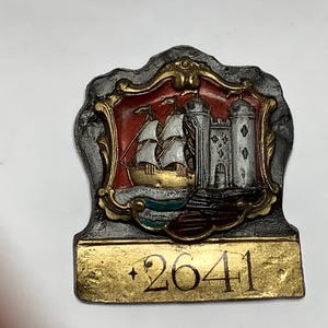 May include: A vintage metal pin featuring a ship and lighthouse scene within a decorative frame. The pin has a gold-colored base with the number "2641" and a plus sign. The ship has white sails and the lighthouse is gray.