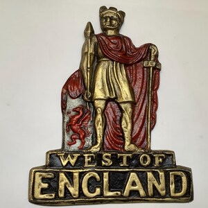 May include: A decorative wall plaque featuring a gold-colored figure of a standing warrior holding a spear and sword, draped in a red cloak. A red dragon is depicted on a shield. The base displays the raised gold text WEST OF ENGLAND on black.