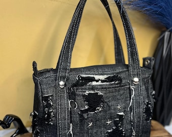 May include: Black denim handbag with distressed details, including rips and star-shaped embellishments. Features two top handles and a front pocket. The bag sits on a wooden surface.
