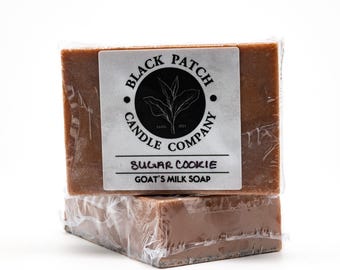 May include: Two bars of brown goat's milk soap, wrapped in clear plastic. Each bar has a white label with the text "BLACK PATCH CANDLE COMPANY" and "SUGAR COOKIE GOAT'S MILK SOAP". The soap is a light brown color.