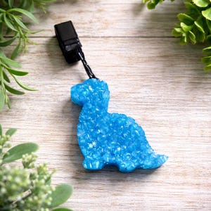 May include: A light blue dinosaur-shaped car air freshener. The air freshener is made of a textured material and has a black clip attached to it. The dinosaur is sitting on a light wood surface.