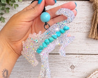 Peut inclure: Un ornement en forme de cheval fabriqué en résine transparente incrustée de paillettes irisées. Le cheval présente une crinière et un dos ornés de petites pierres turquoise irrégulières. Il est suspendu à un cordon noir avec une perle bleu clair.