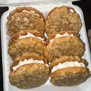May include: Six oatmeal cream pies in a white container. The cookies have a golden-brown exterior with visible oats and a white cream filling. The container is open, revealing the treats.