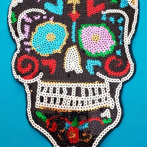 May include: A Day of the Dead-themed skull applique. The skull is black, decorated with colorful sequins in red, blue, yellow, and pink. The skull's outline is white, and the background is a solid teal color.