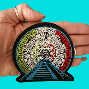 May include: A circular embroidered patch featuring a central sun deity face surrounded by intricate white geometric patterns, with a lime green border on the left, a red border on the right, and a black stepped pyramid silhouette at the base.