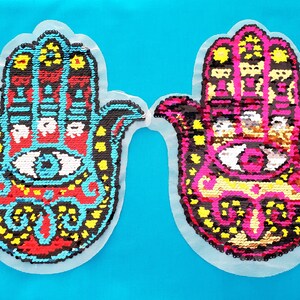 May include: Two colorful Hamsa hand patches with sequins. The Hamsa hands are black, red, yellow, and blue with a white eye in the center. The patches are on a light blue background.