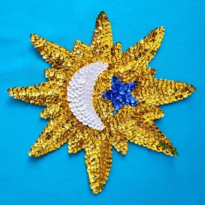 May include: A gold sequin sun with a white crescent moon and a blue star in the center. The sun is on a blue background.
