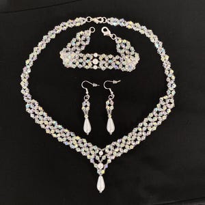 Puede incluir: Un conjunto de collar, pulsera y pendientes de cristal y perlas. El collar tiene un colgante en forma de V con una gran perla colgante. La pulsera y los pendientes están hechos de cuentas de cristal.