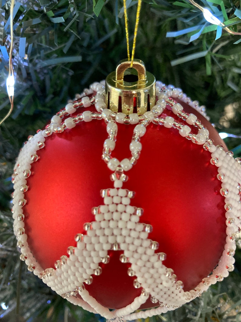 May include: A red Christmas ornament with a white beaded design. The ornament is decorated with a white beaded snowflake pattern.