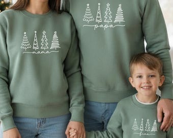 May include: Three people wearing matching sage green sweatshirts with white Christmas tree designs. The sweatshirts have the words "mama", "papa", and "finn" printed below the trees. The family is smiling and holding hands.