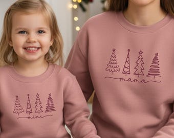 Könnte beinhalten: Zwei passende altrosa Rundhals-Sweatshirts mit einer dunkelroten Grafik aus vier stilisierten Weihnachtsbäumen. Das größere Sweatshirt trägt den Schriftzug mama in Schreibschrift unter den Bäumen, das kleinere mini.