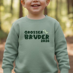 Op de afbeelding: Een saliegroene sweatshirt met de tekst "GROSSER BRUDER 2026" in donkergroen, met een kleine dinosaurusafbeelding. De sweatshirt is gemaakt van een zacht materiaal en is ontworpen voor kinderen.