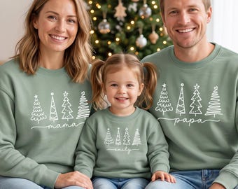 Könnte beinhalten: Drei salbeigrüne Sweatshirts mit Rundhalsausschnitt und weißen Strichzeichnungen von vier stilisierten Weihnachtsbäumen. Die Erwachsenen-Sweatshirts zeigen die Schriftzüge mama und papa, das Kindersweatshirt zeigt emily.