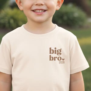Pode incluir: Uma camiseta creme com mangas curtas. A camiseta apresenta o texto "big bro." em marrom, com um coração e o ano "2026" impressos abaixo. A camiseta é projetada para crianças.