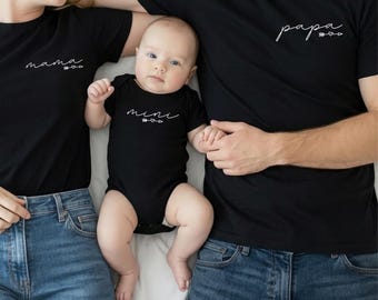 May include: Two black short-sleeved t-shirts and one black short-sleeved baby bodysuit. The left shirt features the white text mama with an arrow and heart. The right shirt features the white text papa with an arrow and heart. The bodysuit features the white text mini with an arrow and heart.
