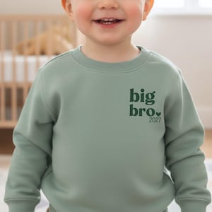 Könnte beinhalten: Ein langärmeliges Sweatshirt in Salbeigrün mit dunkelgrünem Text auf der linken Brust, der big bro. lautet, ergänzt durch ein kleines Herzsymbol und die darunter gedruckte Jahreszahl 2027 in einer klaren, fetten Schriftart.