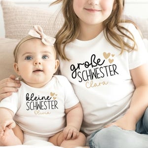 Könnte beinhalten: Ein weißer Kurzarmbody und ein weißes T-Shirt. Der Body zeigt den schwarzen Schriftzug "kleine SCHWESTER" mit "Olivia" darunter und goldene Herzen. Das T-Shirt zeigt den schwarzen Schriftzug "große SCHWESTER" mit "Clara" darunter und goldene Herzen.