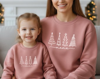 Könnte beinhalten: Zwei altrosa Rundhals-Sweatshirts mit einer weißen Grafik aus vier stilisierten Weihnachtsbäumen. Das größere Sweatshirt trägt den Schriftzug mama in Schreibschrift unter den Bäumen, das kleinere den Schriftzug mini.