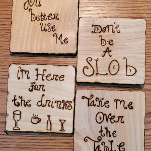 May include: Four square wooden coasters with burnt-in lettering. The coasters have the following phrases: "You better use me", "Don't be a slob", "I'm here for the drinks", and "Take me over the table".