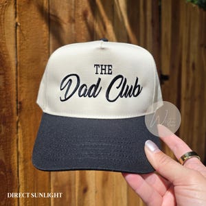 May include: A two-tone baseball cap featuring a cream-colored front panel and a black brim. The front panel displays the embroidered black text "THE" in a block font above the word "Dad Club" in a cursive script font.